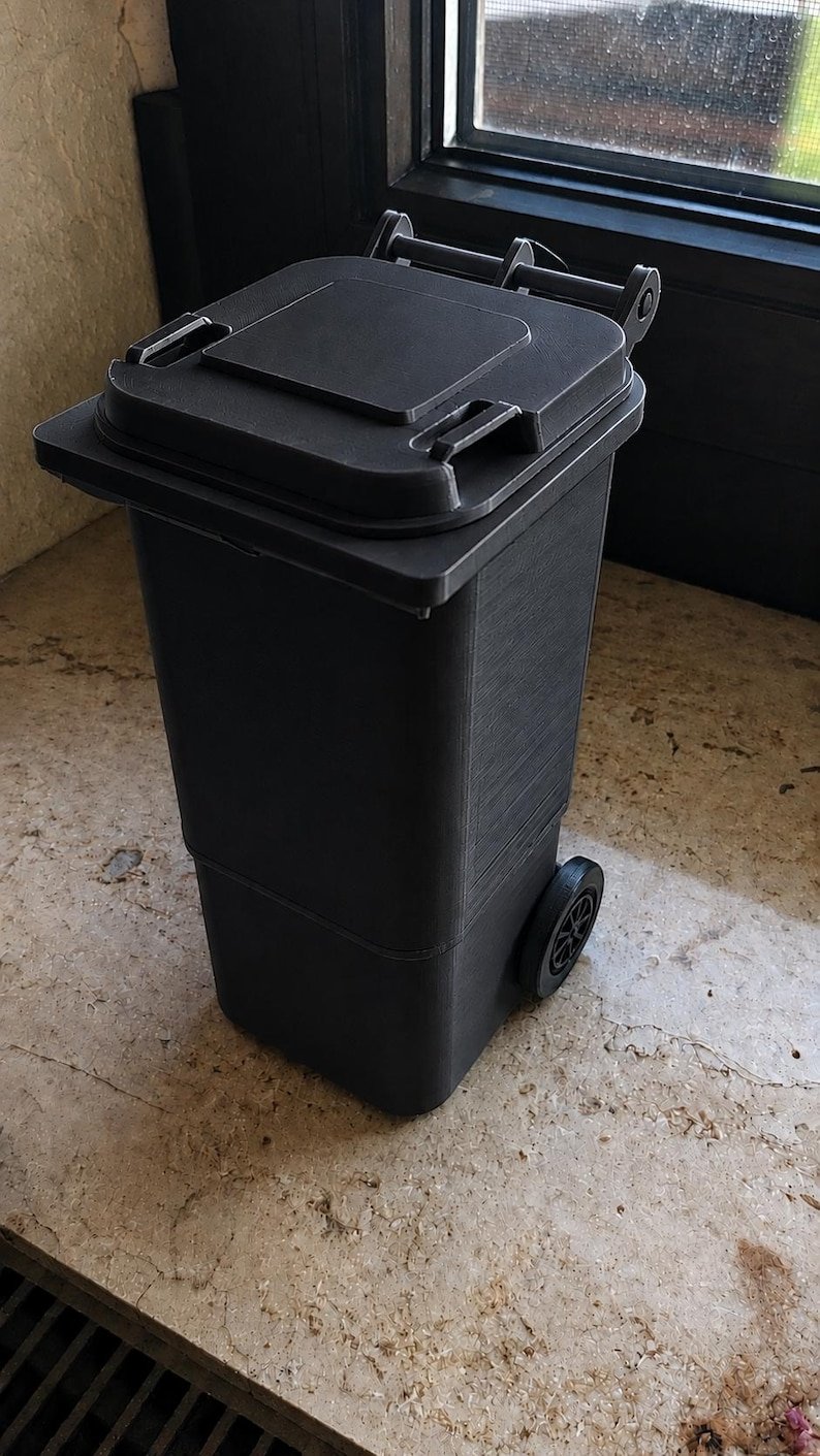Mini Wheelie Bin Trash Can Pencil Holder May include: A small, matte black plastic waste bin featuring a hinged lid with a square centre panel and two side handles. The container is rectangular in shape, stands upright, and is supported by two black wheels located at the lower rear base.
