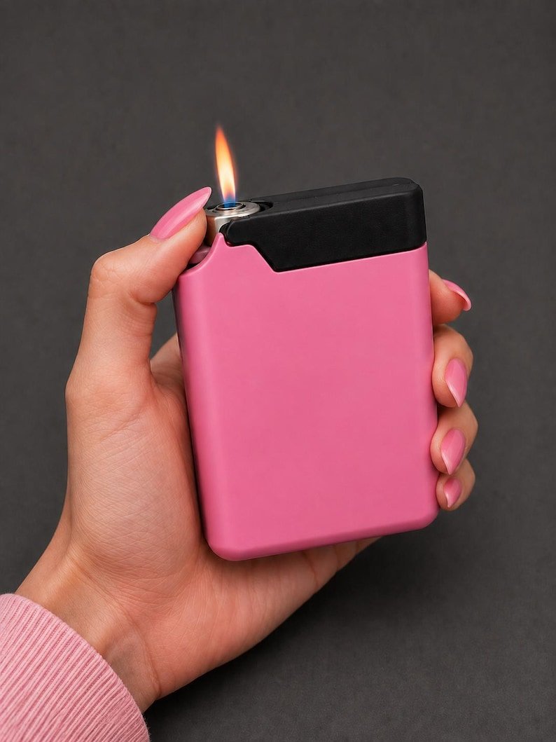 Pink Cigarette Case with Built-In Lighter Holder May include: A rectangular pocket lighter with a matte pink body and a black top section. The lighter features a metallic ignition point emitting a small, visible orange and blue flame. The design is compact with rounded corners and a smooth finish.
