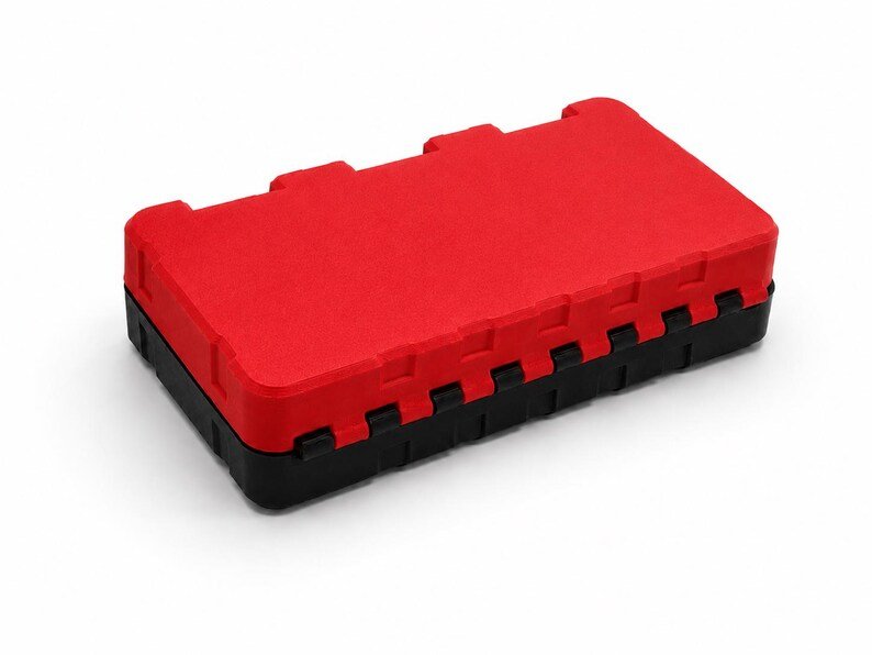 May include: A rectangular storage case featuring a vibrant red lid and a solid black base. The lid has a textured surface with three distinct rectangular indentations along the top edge and a series of interlocking notches along the side.