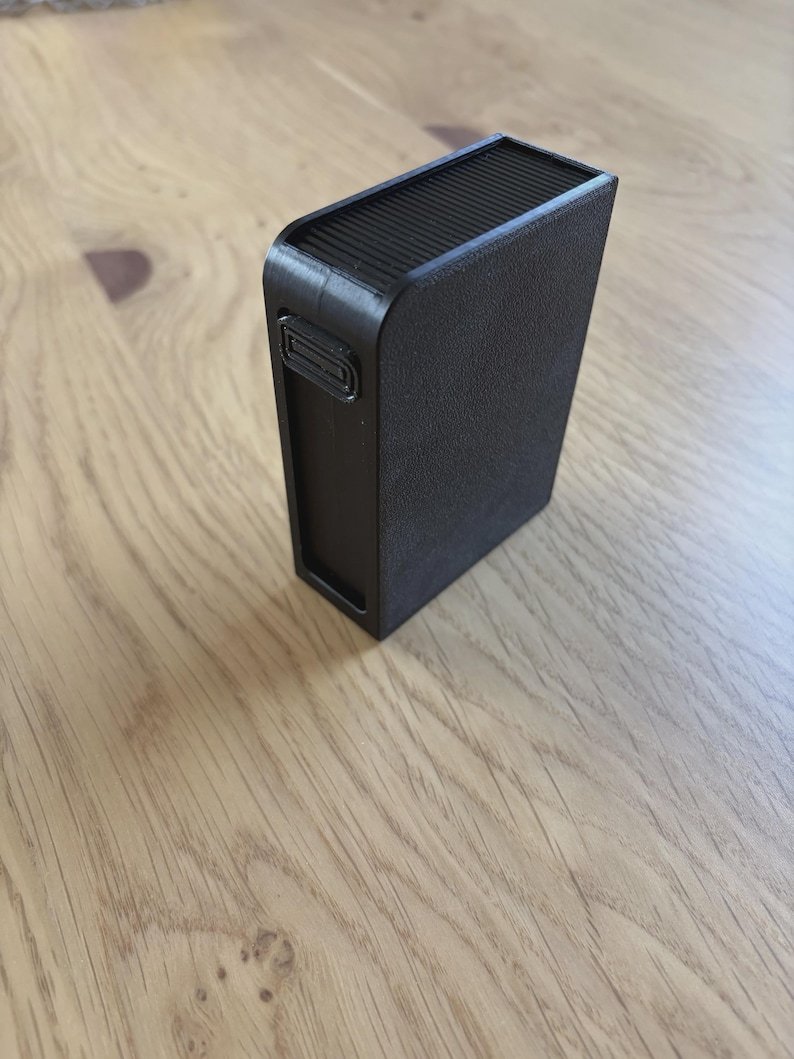 Sliding Cigarette Case — 21 Cigarettes May include: A rectangular black plastic case with a textured finish on the sides and a ribbed top surface. The object features a small, recessed rectangular button on the upper side panel and a smooth, flat front face with a distinct border frame.