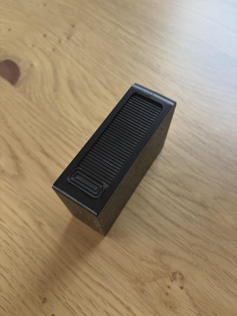 Sliding Cigarette Case — 21 Cigarettes May include: A rectangular black object with a textured surface and a ribbed top panel featuring a small, recessed rectangular button. The device has a matte finish and a structured, boxy shape, showing a top-down angled view of its exterior.
