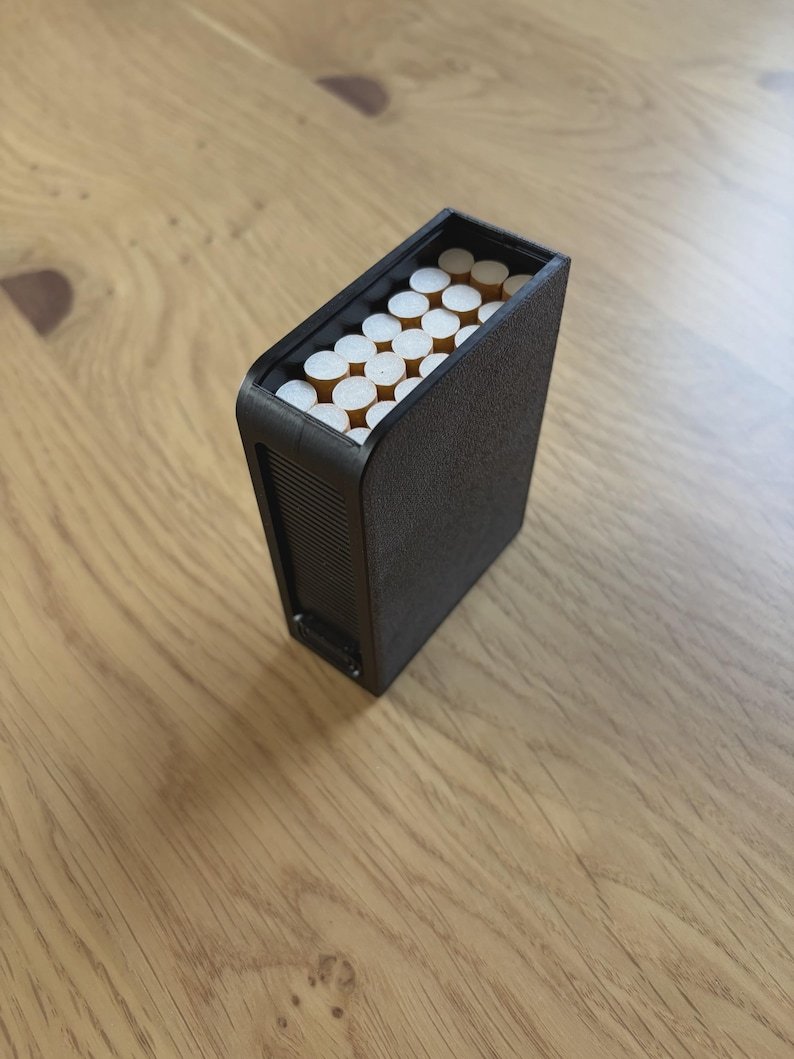 Sliding Cigarette Case — 21 Cigarettes May include: A rectangular black cigarette case with a textured finish, featuring a ribbed side panel and an open top revealing a cluster of white cigarettes with tan filters arranged vertically inside the container.