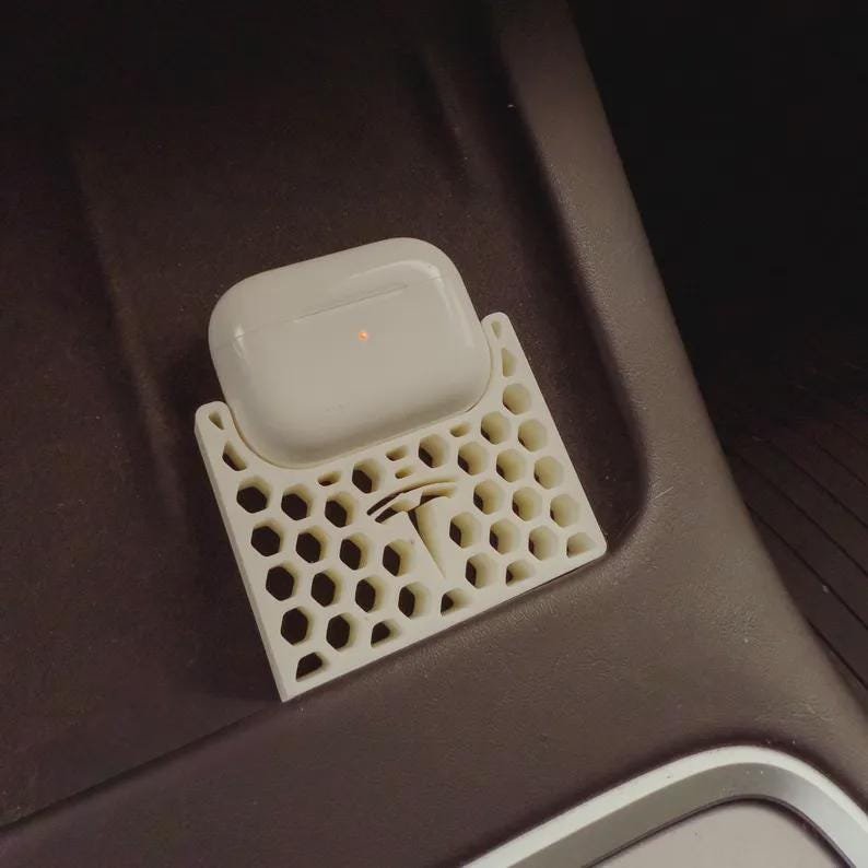 AirPods Pro 2/3 Wireless Charging Spacer for Tesla May include: A white wireless earphone charging case rests on a cream-coloured, 3D-printed plastic tray featuring a hexagonal honeycomb pattern and a central Tesla logo cutout. The tray is designed to fit into a vehicle console storage compartment.