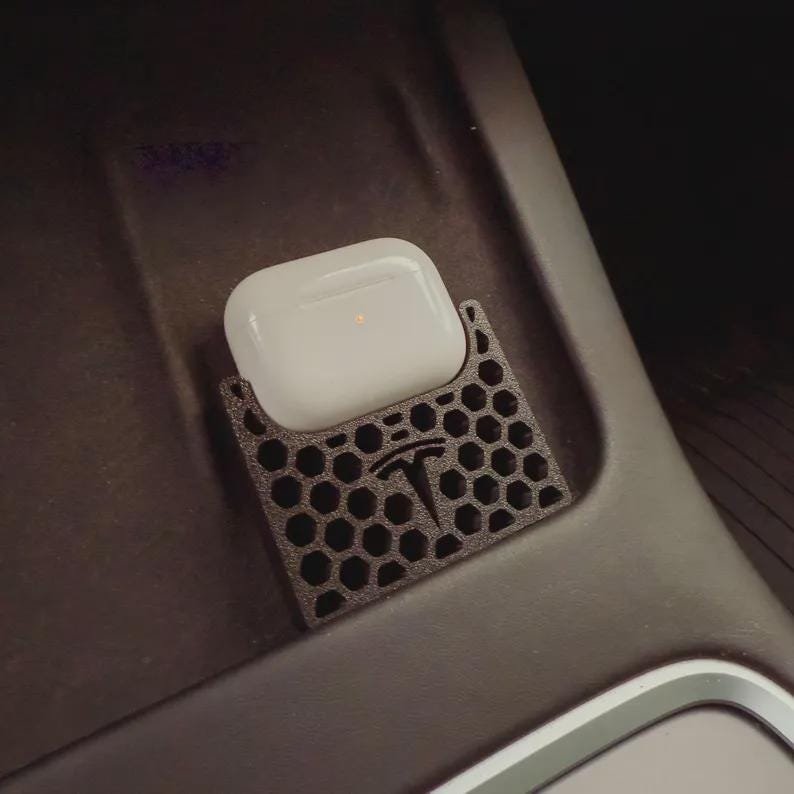 AirPods Pro 2/3 Wireless Charging Spacer for Tesla May include: A white wireless earphone charging case sits inside a black, 3D-printed holder with a hexagonal honeycomb pattern. The holder features a prominent Tesla logo embossed on the front surface, designed to fit into a vehicle console.