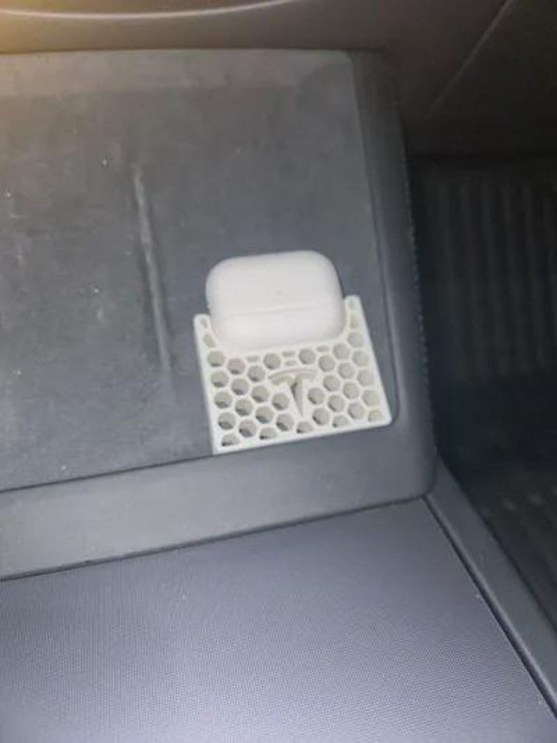 AirPods Pro 2/3 Wireless Charging Spacer for Tesla May include: A white, 3D-printed honeycomb-patterned holder featuring a Tesla logo, designed to secure a white wireless earbud charging case. The accessory is positioned within a dark grey automotive centre console storage area.