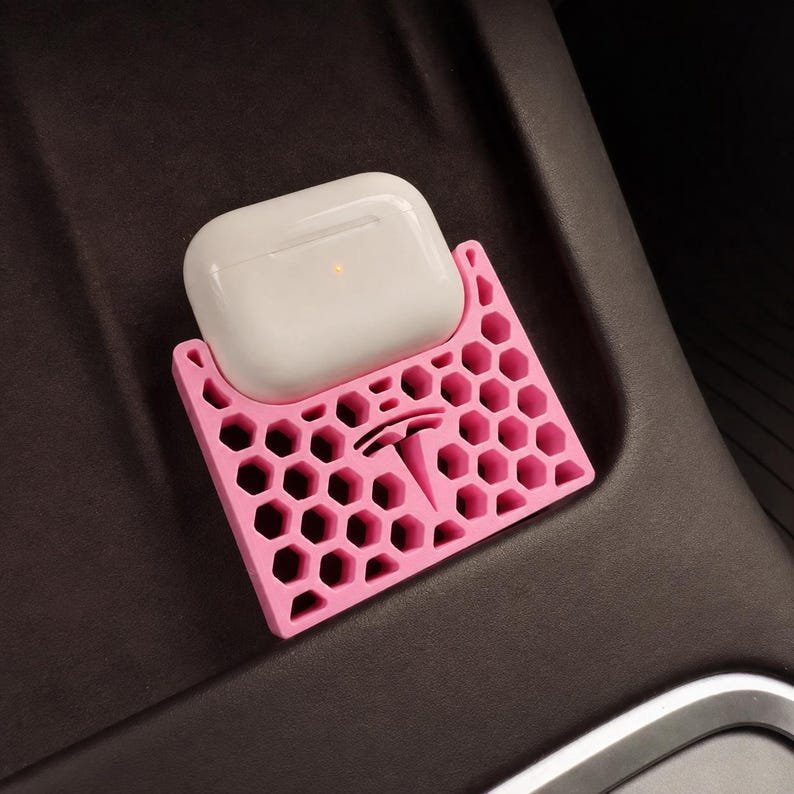 AirPods Pro 2/3 Wireless Charging Spacer for Tesla May include: A pink silicone charging pad featuring a honeycomb pattern and a central Tesla logo. A white wireless earbud charging case with a small orange indicator light is resting on top of the pink accessory, which is designed for a vehicle console.
