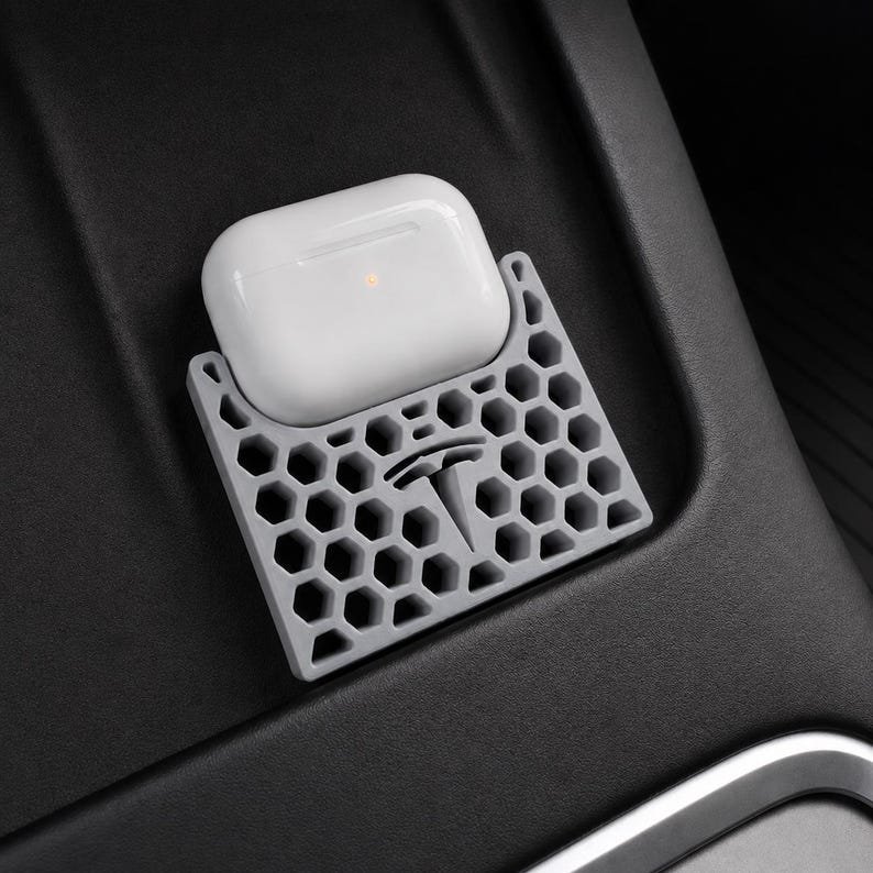 AirPods Pro 2/3 Wireless Charging Spacer for Tesla May include: A white wireless earbud charging case rests inside a grey silicone holder featuring a honeycomb pattern and a Tesla logo cutout. The holder is designed with a rectangular base and open hexagonal cells, providing a secure fit for the white case.