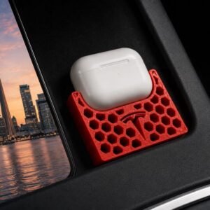 May include: A red 3D-printed honeycomb-patterned storage tray featuring a Tesla logo, designed to hold a white wireless earbud charging case. The tray is positioned within a black automotive console compartment next to a view of a city skyline.