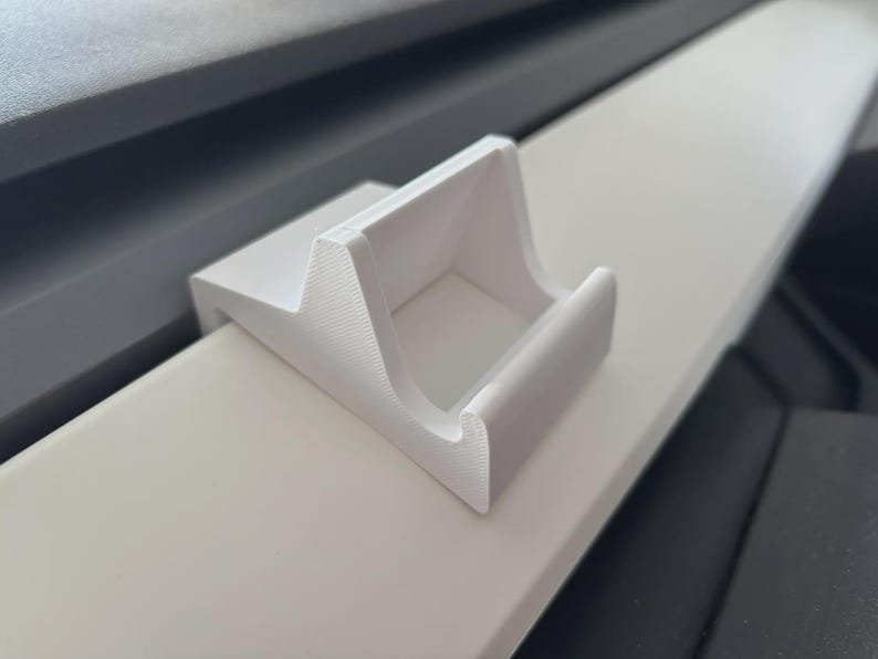 Tesla Model 3/Y Phone Holder May include: A white 3D printed plastic mount with a textured surface and a curved cradle design. The mount features a flat base and a raised front lip, designed to securely hold an object in place. The material shows fine horizontal layer lines.
