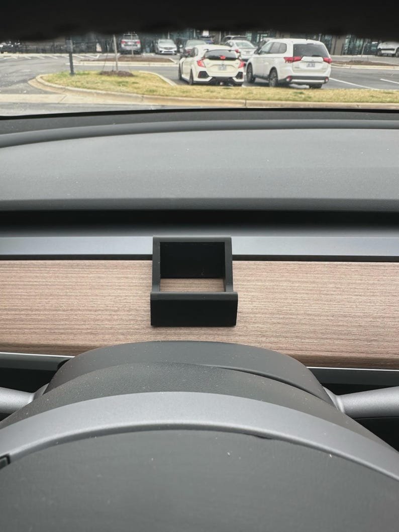 Tesla Model 3/Y Phone Holder May include: A matte black, square-shaped plastic holder mounted onto the wood-grain dashboard trim of a vehicle. The accessory features an open-frame design with a solid base and vertical sides, positioned directly behind the steering wheel column.