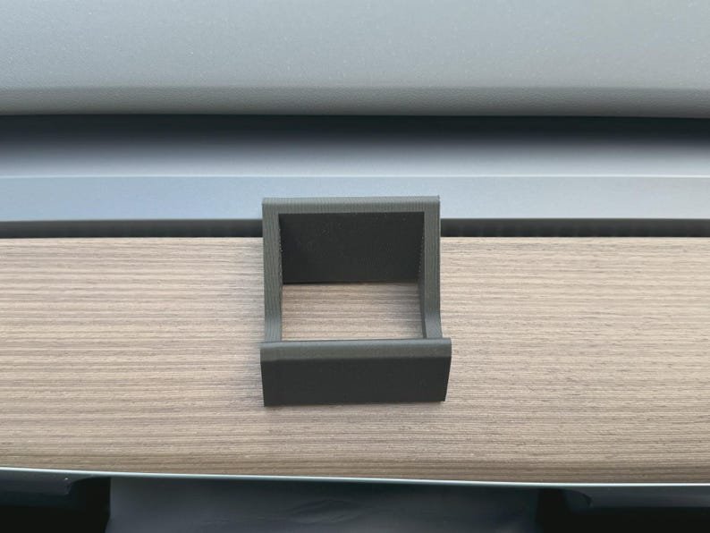 Tesla Model 3/Y Phone Holder May include: A matte black, 3D-printed plastic holder with a square-shaped frame and a front-facing lip, designed to securely hold items in place. The object features a textured surface finish and a hollow centre, providing a stable structure.