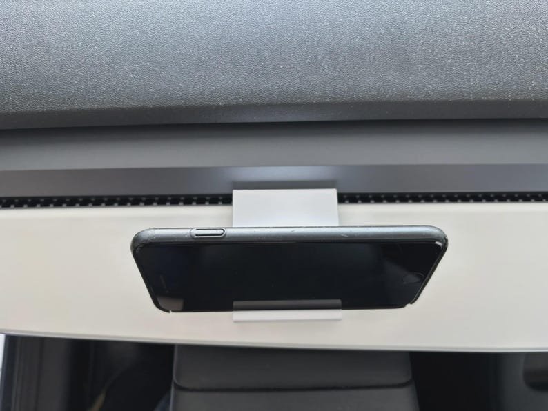 Tesla Model 3/Y Phone Holder May include: A white rectangular phone mount attached to a dashboard, holding a black smartphone in a horizontal position. The mount features a simple, flat design that secures the device against the light-coloured interior panel of the vehicle.
