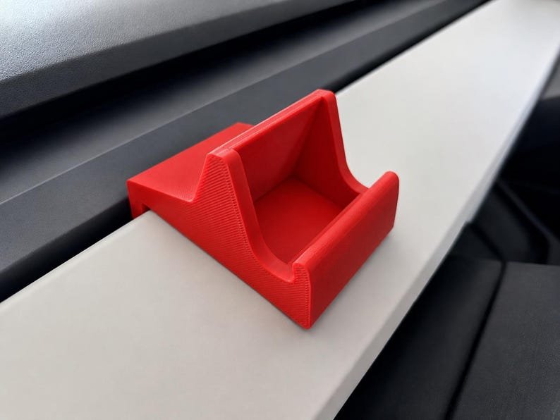 Tesla Model 3/Y Phone Holder May include: A bright red, wedge-shaped phone stand featuring a textured surface and a recessed cradle designed to hold a mobile device securely in place. The stand has a flat base and an angled support structure with a front lip to prevent sliding.