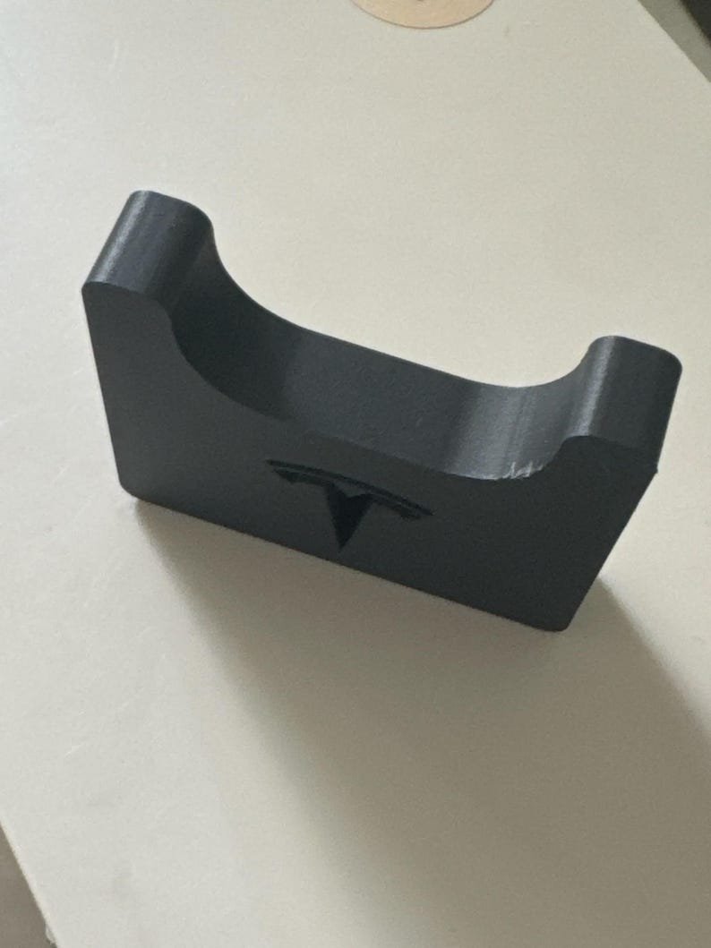 Tesla Model 3 & Model Y Airpods 4 Charging Stand Spacer May include: A matte black rectangular stand featuring a curved top cutout and the Tesla logo embossed on the front face. The object has a solid, block-like construction with smooth edges and a consistent dark finish throughout its entire surface.