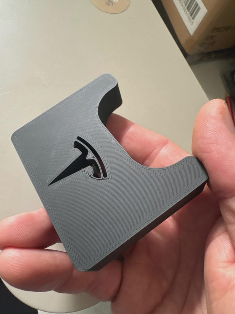Tesla Model 3 & Model Y Airpods 4 Charging Stand Spacer May include: A matte grey 3D printed plastic object with a smooth, flat surface and a curved cutout on one side. The centre of the object features a recessed, glossy black Tesla logo emblem embedded into the grey material with visible print lines.