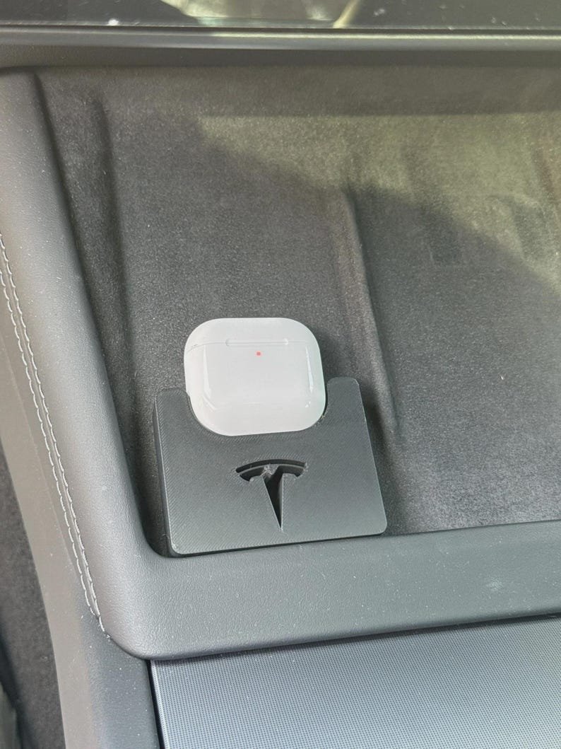 Tesla Model 3 & Model Y Airpods 4 Charging Stand Spacer May include: A white wireless earbud charging case sits inside a black plastic holder featuring a cutout of the Tesla logo. The holder is positioned within a dark grey automotive centre console storage area, with a small red light visible on the case.