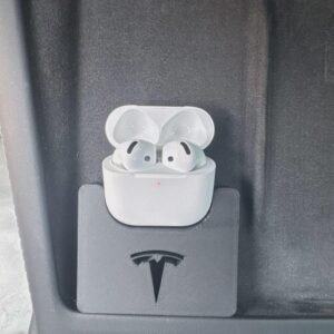 May include: A white wireless earbud charging case with two earbuds inside, featuring a small orange status light. The case is held in a black, rectangular, 3D-printed plastic holder that displays a cutout of the Tesla logo on its front surface.