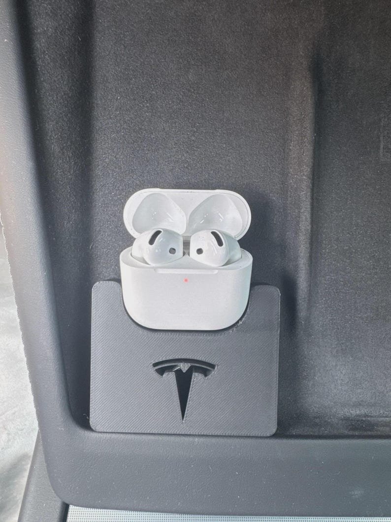 Tesla Model 3 & Model Y Airpods 4 Charging Stand Spacer May include: A white wireless earbud charging case with two earbuds inside, featuring a small orange status light. The case is held in a black, rectangular, 3D-printed plastic holder that displays a cutout of the Tesla logo on its front surface.