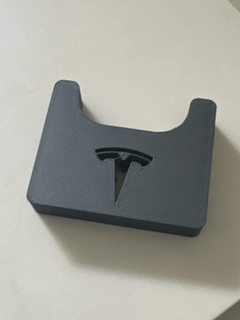 Tesla Model 3 & Model Y Airpods 4 Charging Stand Spacer May include: A dark grey, rectangular, 3D-printed plastic holder featuring a U-shaped cutout on the top edge and a recessed Tesla logo centred on the front face. The surface exhibits a visible diagonal layer line texture from the printing process.