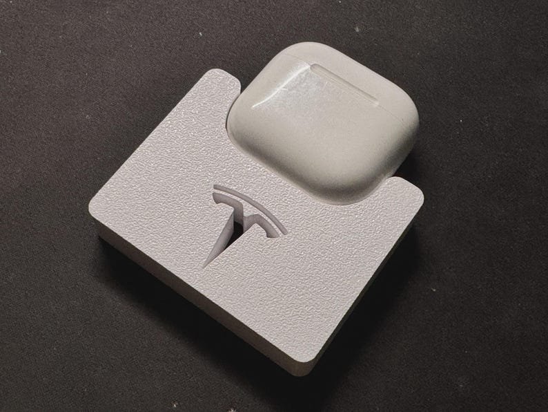 Tesla Model 3 & Model Y Airpods 4 Charging Stand Spacer May include: A white, textured, square-shaped charging dock featuring a cutout of the Tesla logo. The dock is designed to hold a white, glossy, rectangular wireless earbud charging case, which is currently seated in the top section of the stand.
