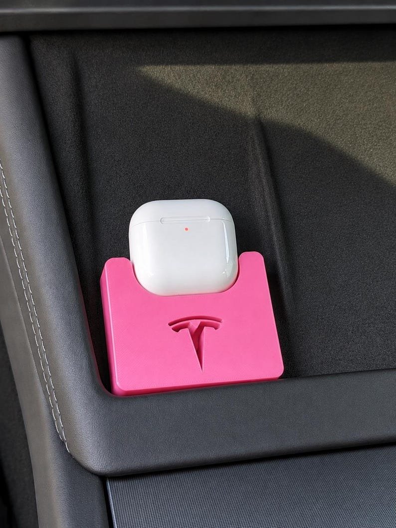 Tesla Model 3 & Model Y Airpods 4 Charging Stand Spacer May include: A bright pink plastic holder featuring a debossed Tesla logo, designed to securely store a white wireless earbud charging case. The holder is positioned within a dark grey interior console, providing a dedicated space for the device.