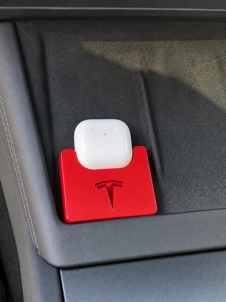 Tesla Model 3 & Model Y Airpods 4 Charging Stand Spacer May include: A bright red plastic holder featuring a debossed Tesla logo, designed to secure a white wireless earbud charging case. The holder is positioned on a dark grey textured surface, providing a stable storage solution for small electronics.