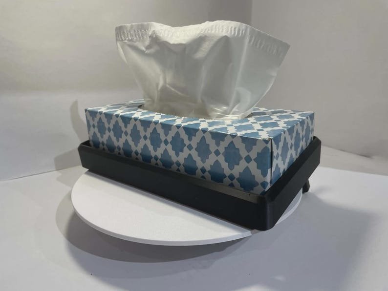 Tesla Model 3 & Model Y Tissue Box Holder May include: A rectangular black tissue box holder containing a cardboard box with a blue and white geometric lattice pattern. A single white facial tissue is pulled through the top opening of the box, extending upwards from the container.