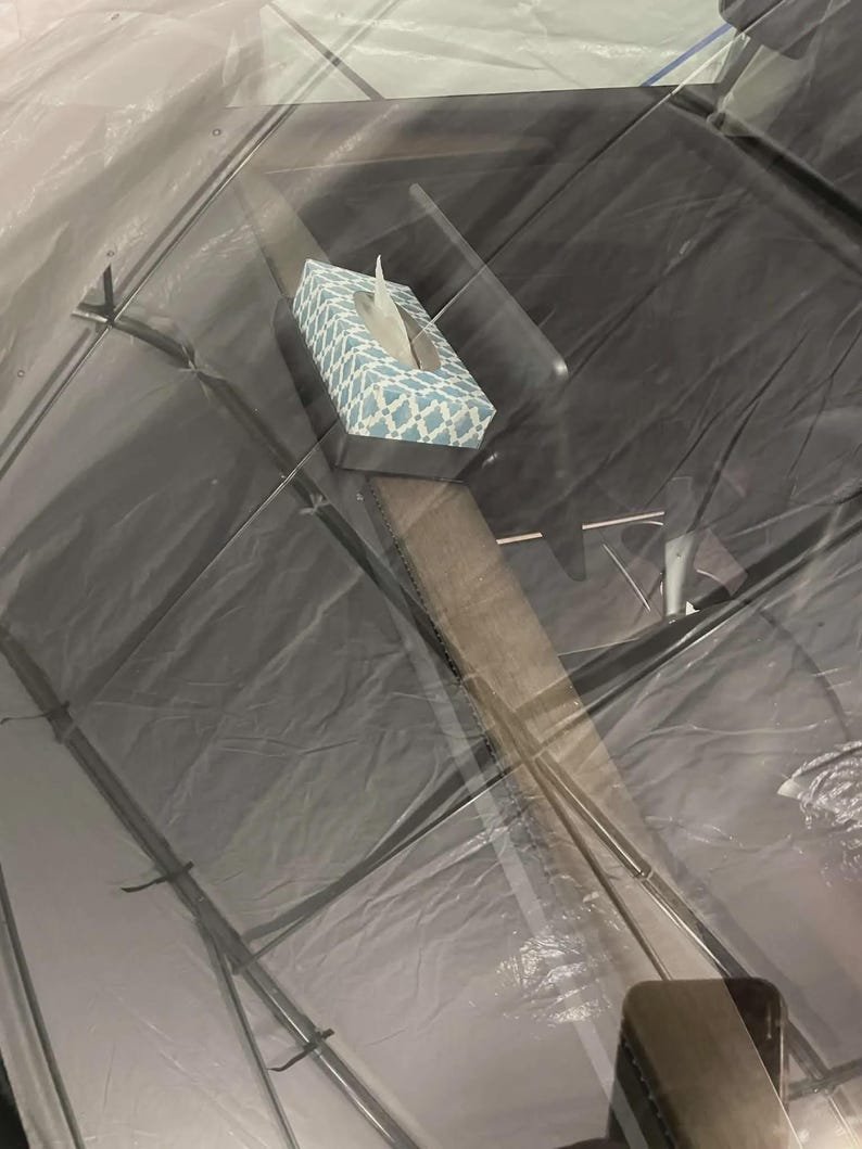 Tesla Model 3 & Model Y Tissue Box Holder May include: A rectangular tissue box holder featuring a light blue and white geometric lattice pattern. The box is positioned on a dark surface, viewed through a transparent, slightly wrinkled plastic covering that creates reflections and distortions.