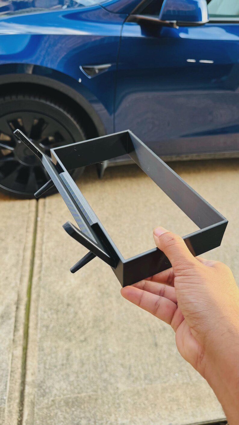 Tesla Model 3 & Model Y Tissue Box Holder May include: A black rectangular plastic frame with a protruding angled support arm and a small perpendicular peg on one side. The object features a hollow centre and a smooth, matte finish, designed with a rigid structure and clean, sharp edges.