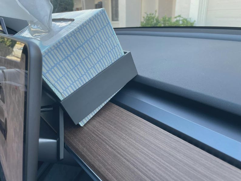 Tesla Model 3 & Model Y Tissue Box Holder May include: A dark grey plastic tissue box holder mounted to the side of a vehicle dashboard display screen. The holder contains a rectangular tissue box featuring a light blue and white grid pattern with tissues protruding from the top opening.