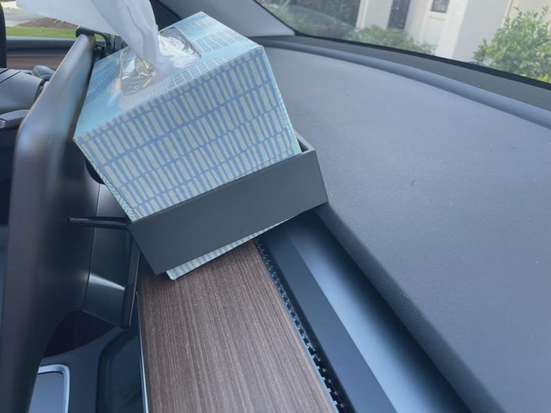 Tesla Model 3 & Model Y Tissue Box Holder May include: A rectangular dark grey plastic tissue box holder mounted to the back of a vehicle display screen. The holder contains a light blue and white patterned cardboard tissue box with a white tissue protruding from the top opening.