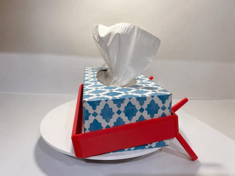 Tesla Model 3 & Model Y Tissue Box Holder May include: A rectangular tissue box with a blue and white geometric pattern, containing white tissues, held within a bright red plastic frame. The frame features angled support legs extending outward from the sides, providing a stable base.