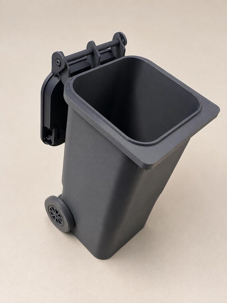 Mini Wheelie Bin Trash Can Pencil Holder May include: A dark grey plastic waste bin featuring a hinged lid that is open and positioned at the back. The container has a rectangular body with a wide top opening and two small wheels attached to the lower sides for mobility.