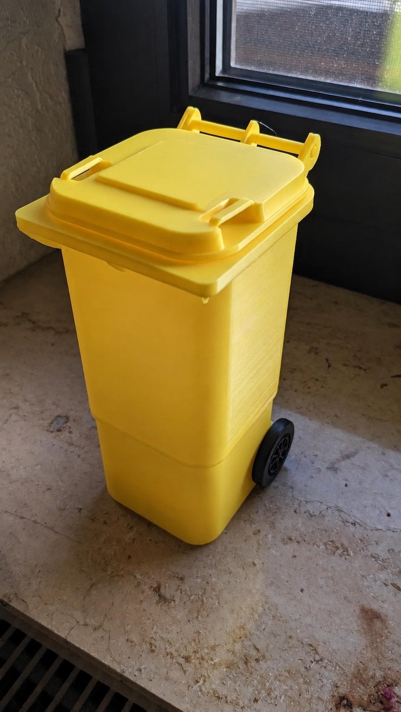 Mini Wheelie Bin Trash Can Pencil Holder May include: A bright yellow miniature waste bin featuring a hinged lid and two small black wheels on the sides. The container has a rectangular body with a slightly tapered base and a textured surface finish, resembling a standard outdoor wheelie bin.