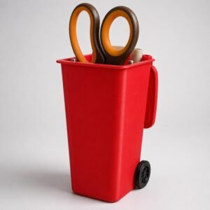 May include: A bright red desk organiser shaped like a miniature rolling bin with a hinged lid and two small black wheels at the base. The container holds a pair of scissors with brown and orange handles and a small beige cylindrical object.