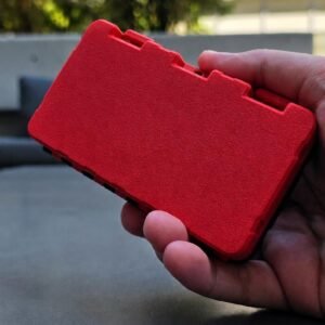 May include: A rectangular storage case with a textured bright red exterior and a black base. The case features a hinged lid with a notched edge design along the top side, providing a secure closure mechanism for the internal compartment.