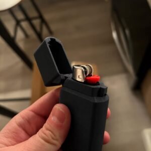 May include: A matte dark grey rectangular lighter case with a hinged lid in the open position. The case holds a standard metal lighter insert featuring a silver-coloured ignition wheel and a bright red plastic fuel release button on the side.