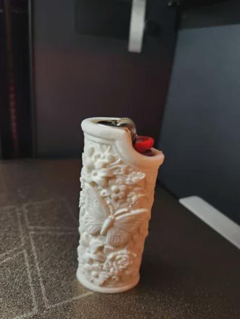 Butterfly Garden Lighter Sleeve May include: A white lighter featuring a detailed, raised relief texture of butterflies and various floral patterns covering the entire exterior surface. The top of the lighter includes a metallic ignition wheel and a small, bright red plastic button.