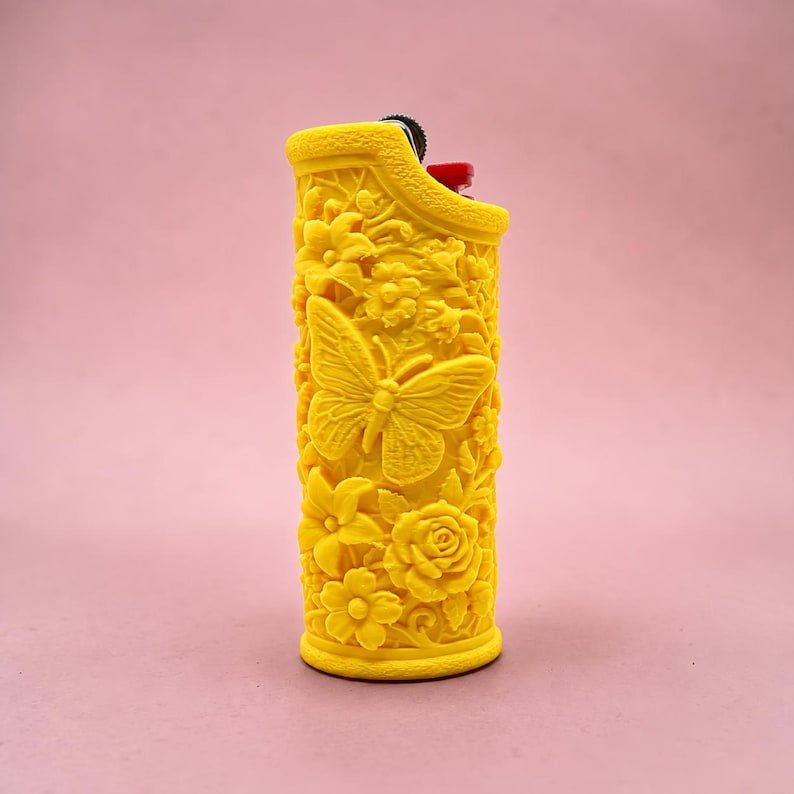 Butterfly Garden Lighter Sleeve May include: A bright yellow lighter featuring a detailed, raised relief design of a butterfly surrounded by various flowers and foliage. The top of the lighter shows a small black ignition wheel and a red plastic button for operation.