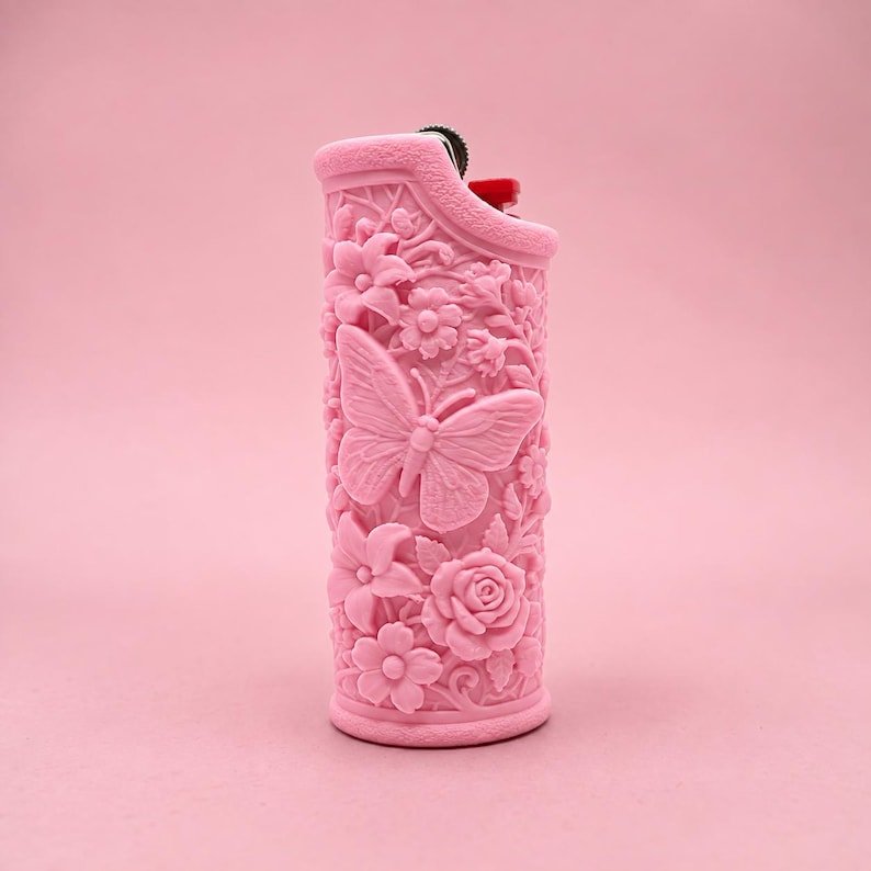 Butterfly Garden Lighter Sleeve May include: A light pink lighter featuring a raised, textured relief design of a central butterfly surrounded by various flowers and foliage. The lighter has a metallic ignition mechanism at the top with a small red component visible.