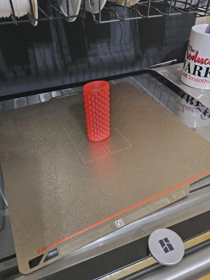 Smooth Dragon Scale BIC Lighter Sleeve May include: A cylindrical, red, 3D-printed object with a textured, spiked surface. The object is positioned on a flat, metallic, square-shaped build plate with a visible red line along the bottom edge and a small square logo near the centre.
