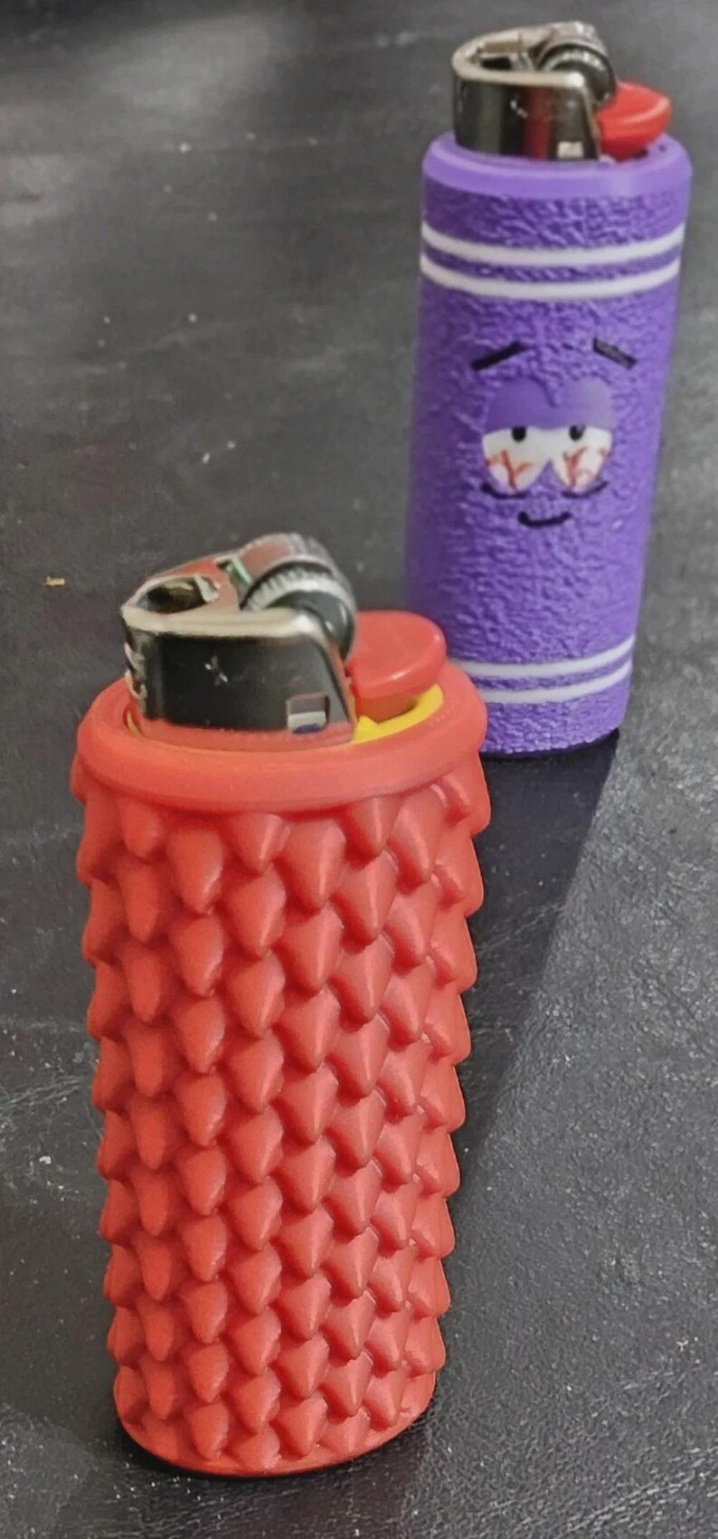 Smooth Dragon Scale BIC Lighter Sleeve May include: Two lighters with protective silicone sleeves. The front lighter is red with a textured, spiked surface. The rear lighter is purple with a textured finish, white horizontal stripes, and a graphic of a face with bloodshot eyes.