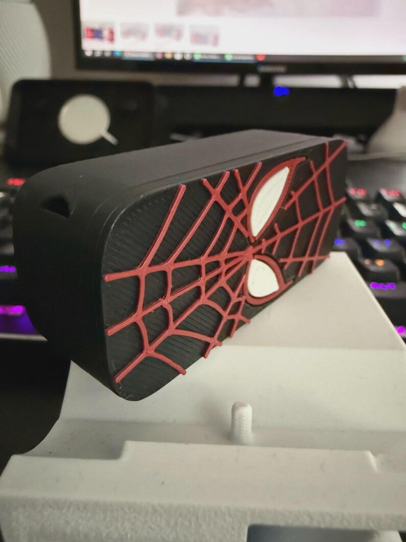 Spider-Inspired Lighter Case May include: A rectangular black protective case featuring a raised red spiderweb pattern and two white eye-shaped accents on the front surface. The case has a smooth matte finish and a small circular cutout on the side edge for cable access.