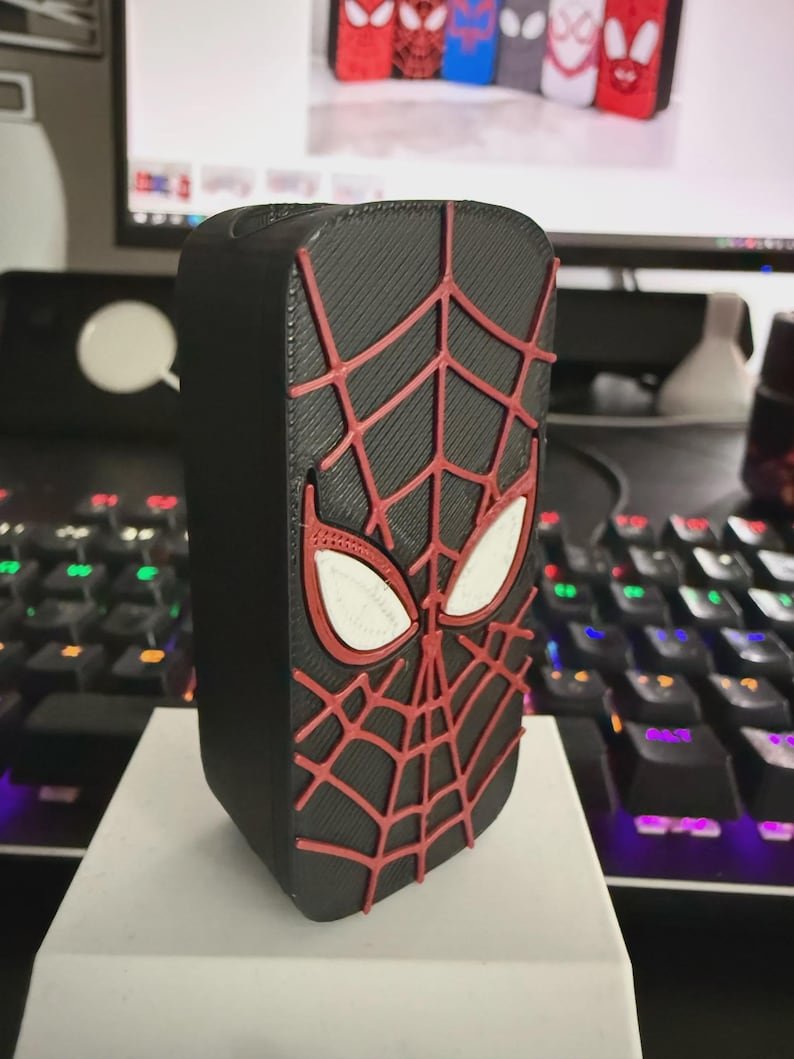 Spider-Inspired Lighter Case May include: A rectangular black plastic case featuring a raised red spiderweb pattern and two white eye-shaped accents on the front. The design mimics a superhero mask with a textured surface, showcasing a detailed web structure across the black base.