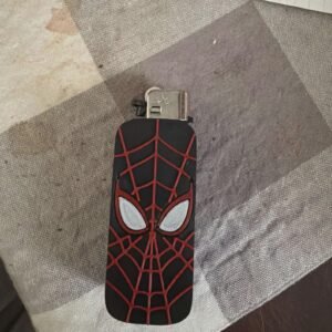 Home May include: A black rectangular lighter case featuring a raised red spiderweb pattern and two white eye-shaped accents. The metallic ignition mechanism is visible at the top of the case, which is designed to fit over a standard lighter.