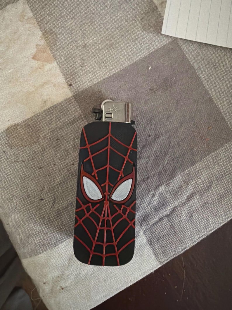 Spider-Inspired Lighter Case May include: A black rectangular lighter case featuring a raised red spiderweb pattern and two white eye-shaped accents. The metallic ignition mechanism is visible at the top of the case, which is designed to fit over a standard lighter.