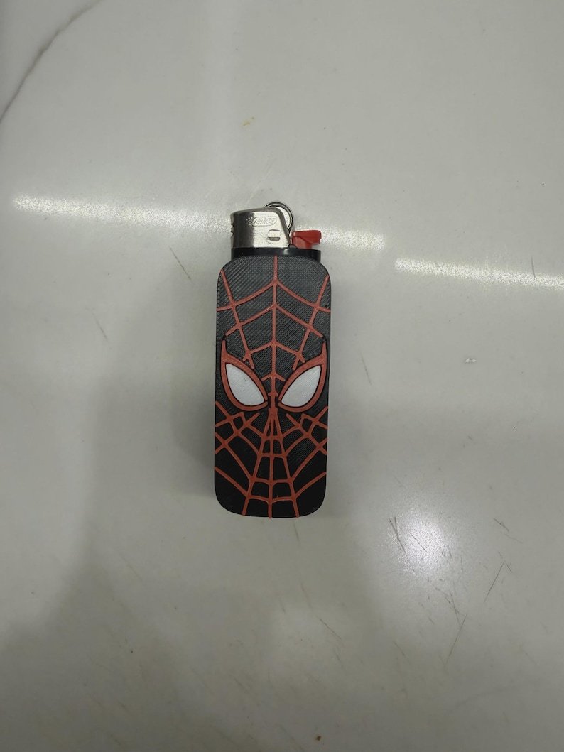 Spider-Inspired Lighter Case May include: A rectangular pocket lighter featuring a black textured surface with a red spiderweb pattern and two white eye-shaped graphics. The top of the lighter is metallic silver with a small red ignition button and a metal loop for attachment.