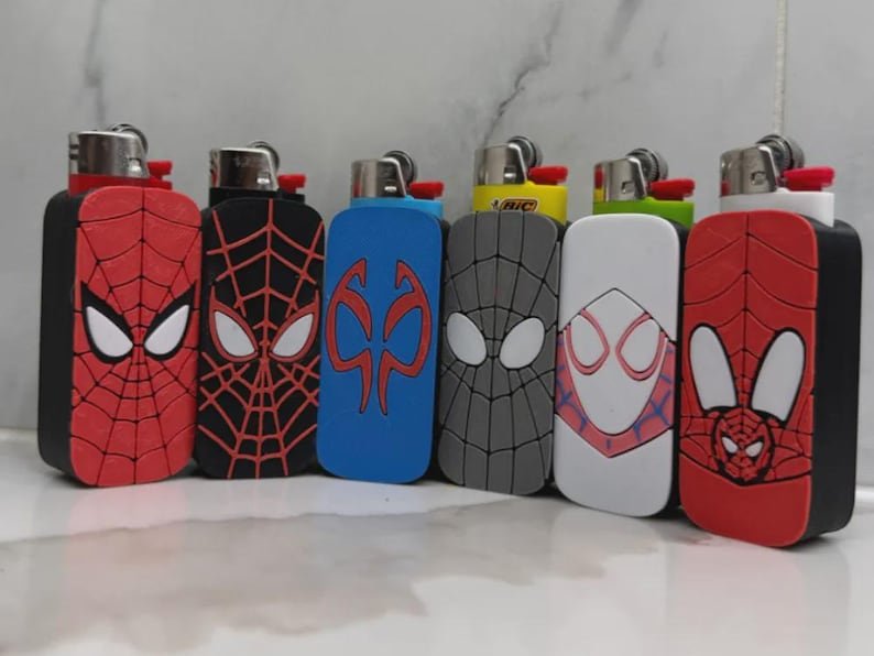 Spider-Inspired Lighter Case May include: Six rectangular lighter cases featuring various superhero-inspired designs, including red and black spider-web patterns, a blue emblem, a grey webbed mask, and a white mask design, each fitted with a standard metal-topped lighter.