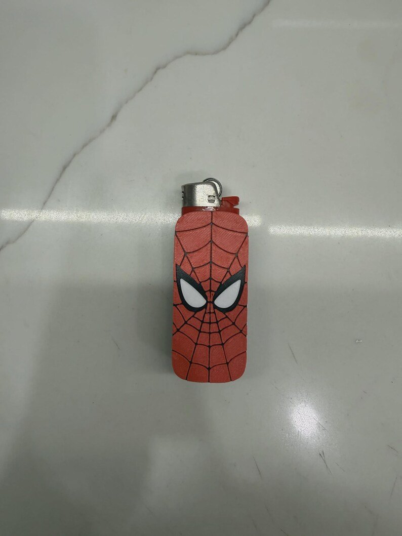 Spider-Inspired Lighter Case May include: A rectangular lighter featuring a red exterior with a black web pattern and two large white eyes with black outlines. The top of the lighter has a metallic silver ignition mechanism and a red plastic base component.