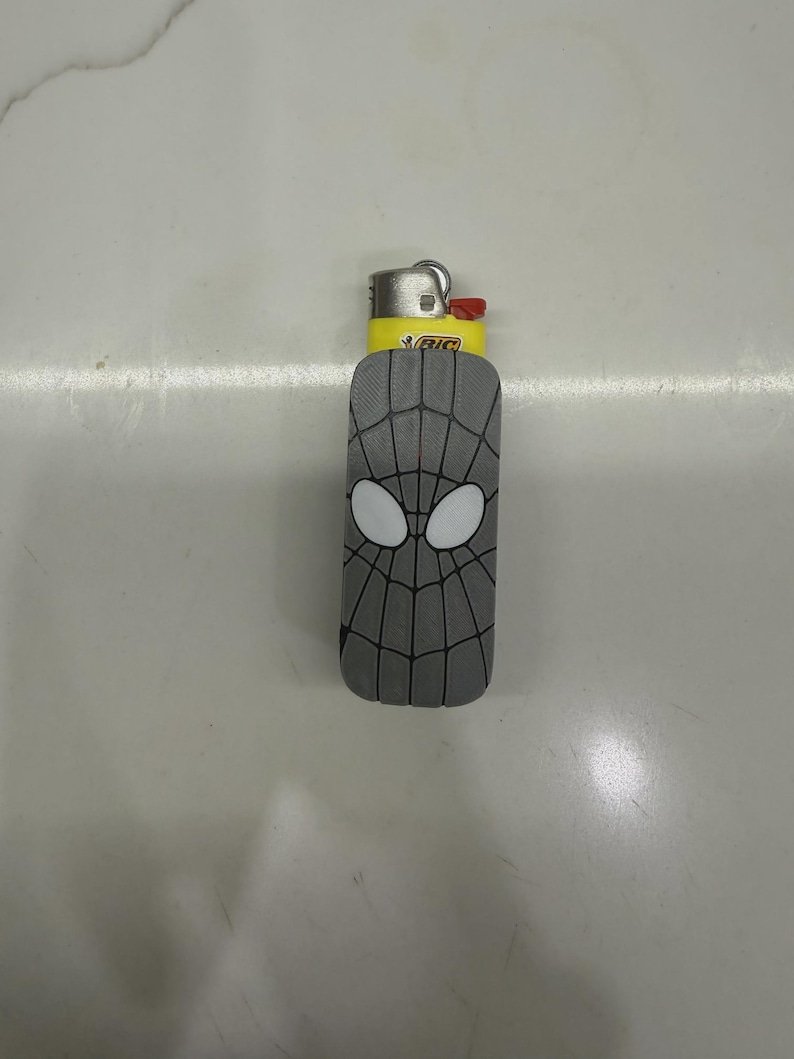 Spider-Inspired Lighter Case May include: A yellow BIC lighter encased in a grey sleeve featuring a black web pattern and two white oval eye shapes. The lighter has a metallic top with a red ignition button and the BIC logo printed in black on a yellow background.